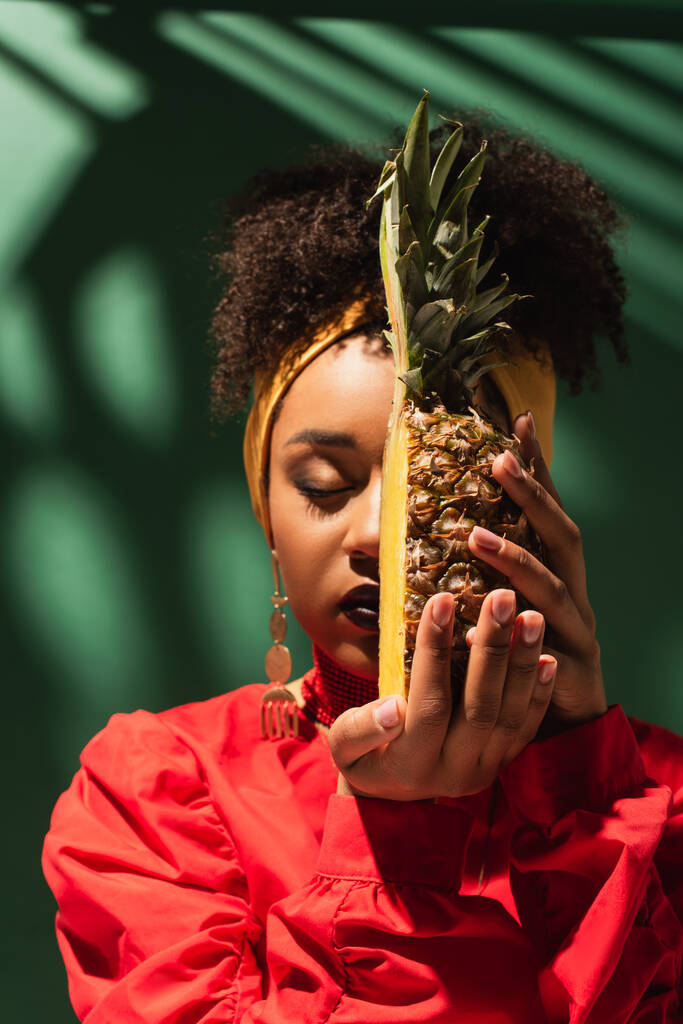 young african american woman holding half cut of pineapple near face on green - Photo, Image