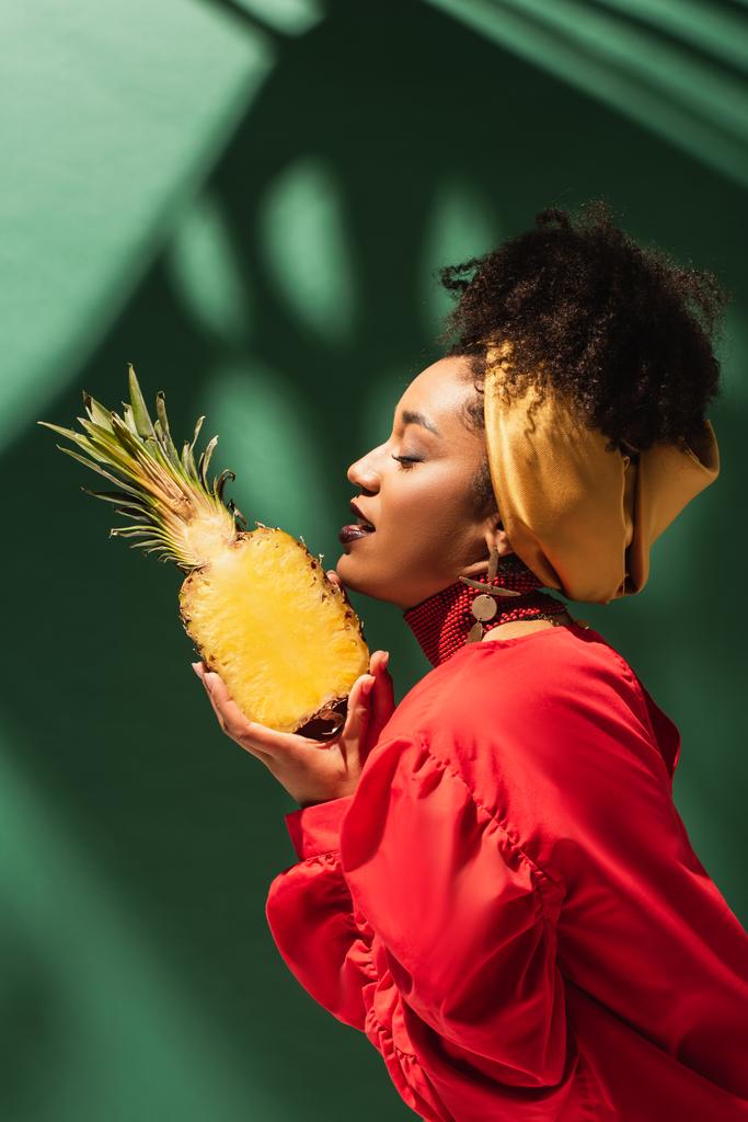 vue latérale de la jeune femme afro-américaine avec les yeux fermés tenant demi-coupe d'ananas dans les mains sur vert - Photo, image