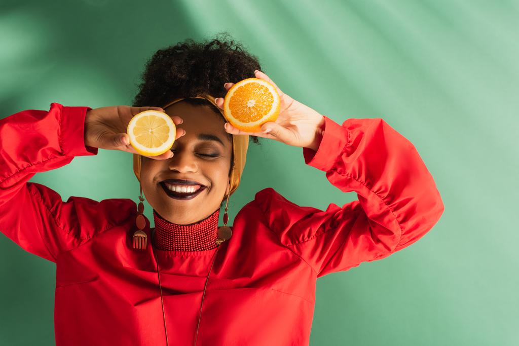 улыбающаяся молодая африканская американка, держащая половину лимона и оранжевого рядом с лицом на зеленом - Фото, изображение