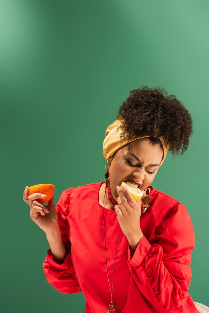giovane donna afroamericana in possesso di arancione e mangiare mezzo taglio di limone sul verde - Foto, immagini