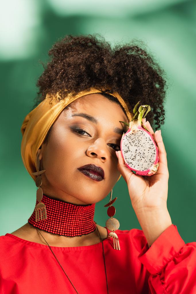 νεαρή Αφροαμερικανή γυναίκα με μαντίλα που κρατάει μισό κομμένο φρούτο δράκου σε πράσινο. - Φωτογραφία, εικόνα