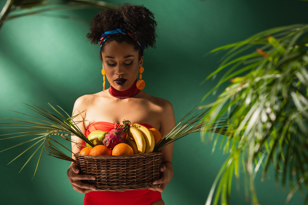 молодая африканская американка смотрит на корзину с экзотическими фруктами на зеленом  - Фото, изображение