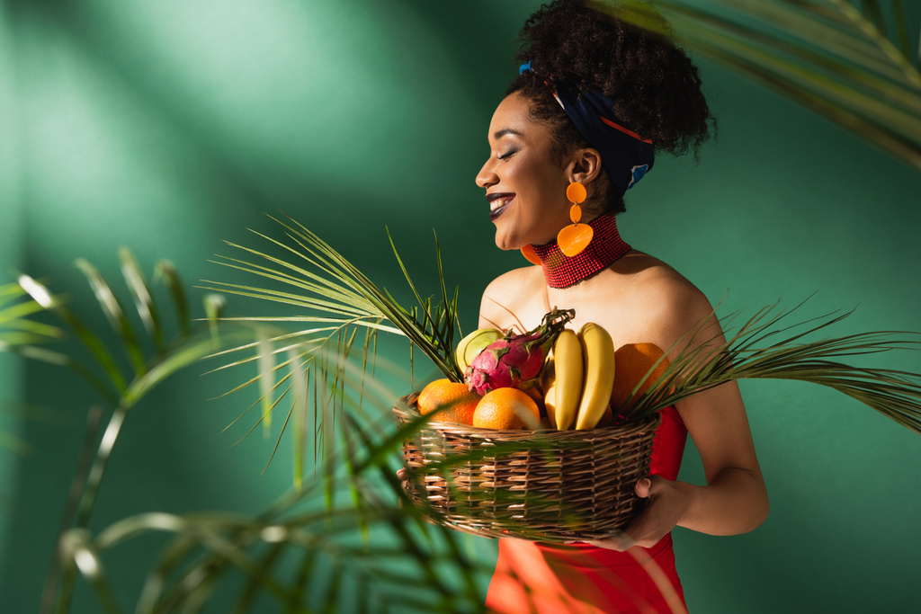 smiling young african american woman in red swimsuit holding basket with exotic fruits on green - Photo, Image