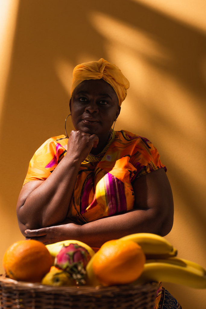 figyelmes középkorú afro-amerikai eladónő ül gyümölcsök közelében, kezét közel arc narancs - Fotó, kép