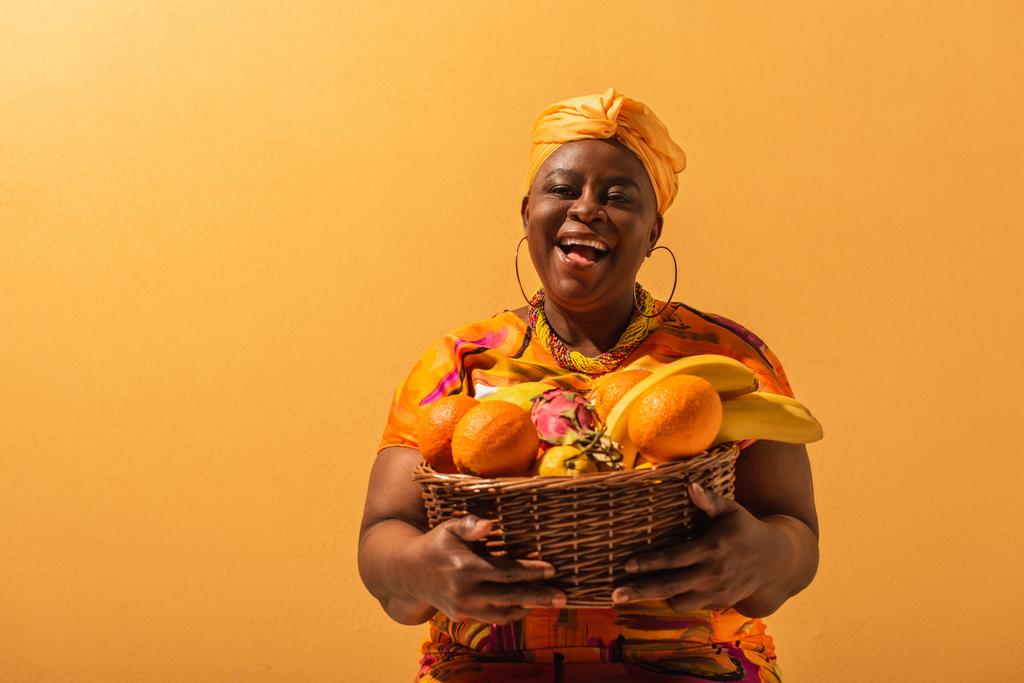 sourire femme afro-américaine d'âge moyen tenant panier avec des fruits sur orange - Photo, image