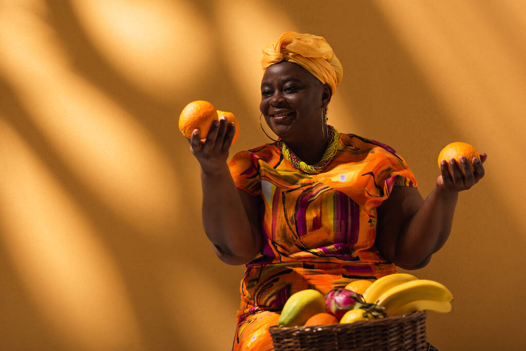 pozytywna afrykańska sprzedawczyni w średnim wieku trzymająca pomarańcze w rękach i sprzedająca owoce w tle z cieniami - Zdjęcie, obraz