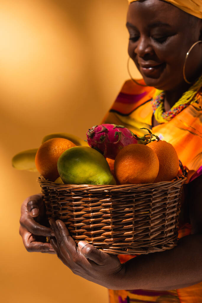 Африканская женщина средних лет в ярком платье держит корзину с экзотическими фруктами на апельсине - Фото, изображение