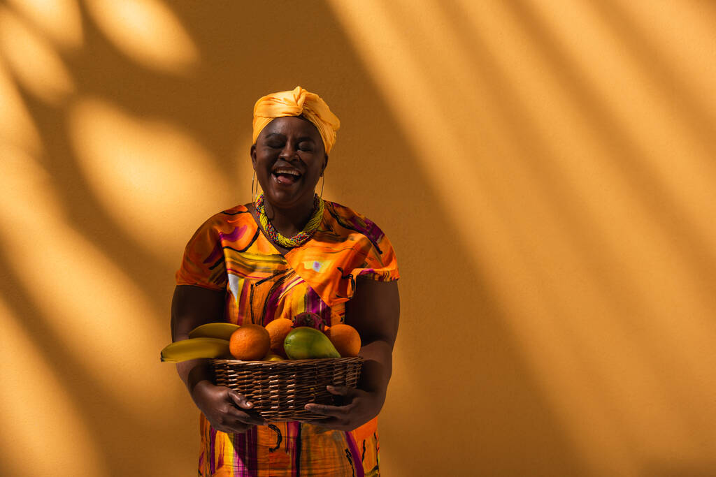 smiling middle aged african american woman in bright dress and turban holding basket with exotic fruits on orange - Photo, Image
