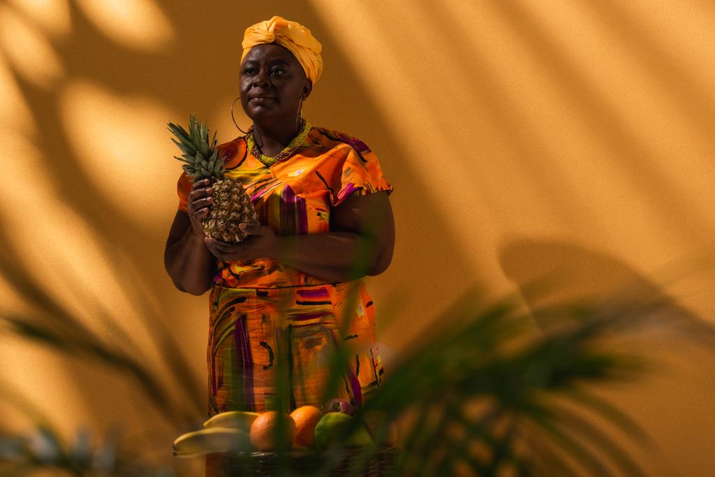 Afrikanische Verkäuferin mittleren Alters steht mit Ananas in der Nähe von Früchten auf Orange - Foto, Bild