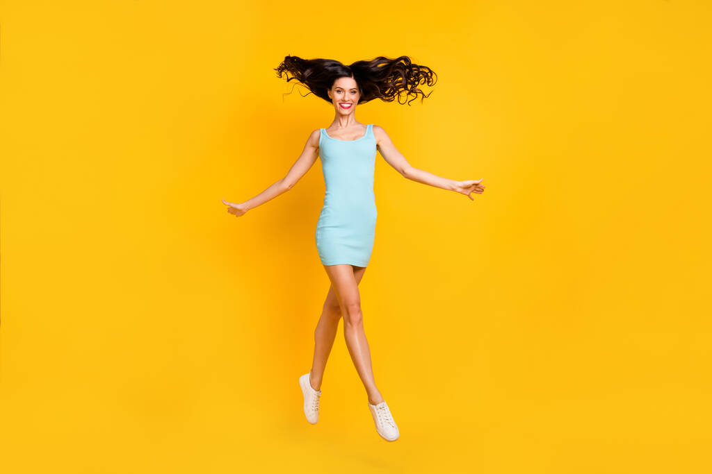 Φωτογραφία από γοητευτικό νεαρή κοπέλα άλμα λαμπερό χαμόγελο άνεμος φυσήξει τα μαλλιά φορούν μπλε κοντά παπούτσια φόρεμα απομονωμένο κίτρινο χρώμα φόντο - Φωτογραφία, εικόνα