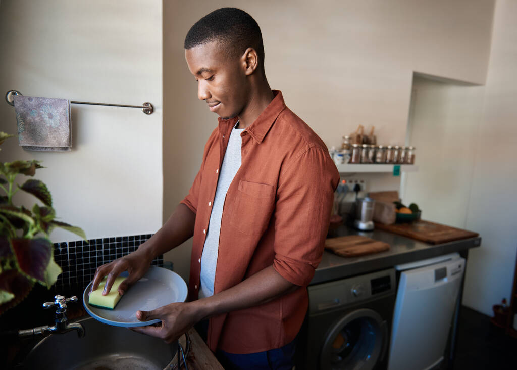 Молодой африканский мужчина моет посуду в раковине на кухне, выполняя работу вокруг своей квартиры - Фото, изображение