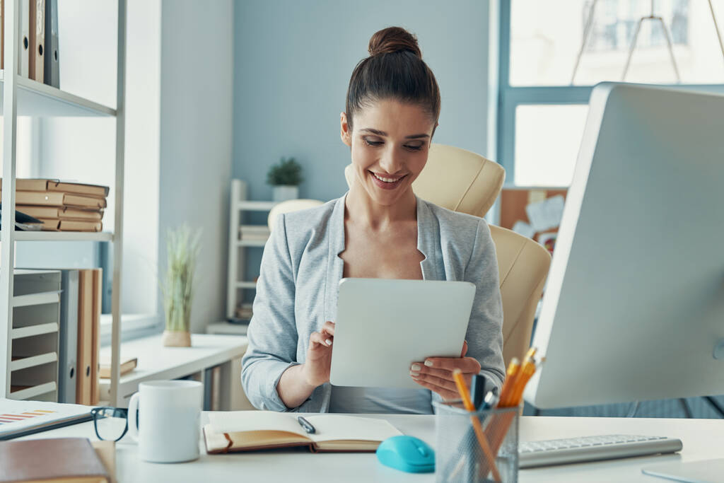 красивая молодая женщина в элегантной повседневной одежде с помощью цифрового планшета и улыбаясь, сидя в офисе - Фото, изображение