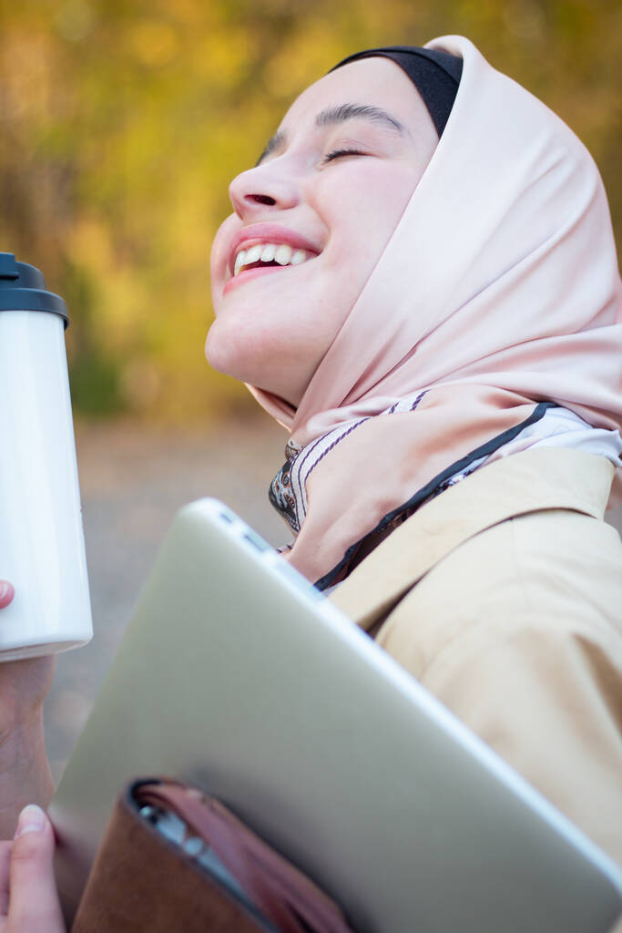 Muzułmanka w hidżabie w beżowym płaszczu z filiżanką kawy i laptopem w parku. Muzułmańska bizneswoman. Uczeń. Bliski plan, twarz.. - Zdjęcie, obraz