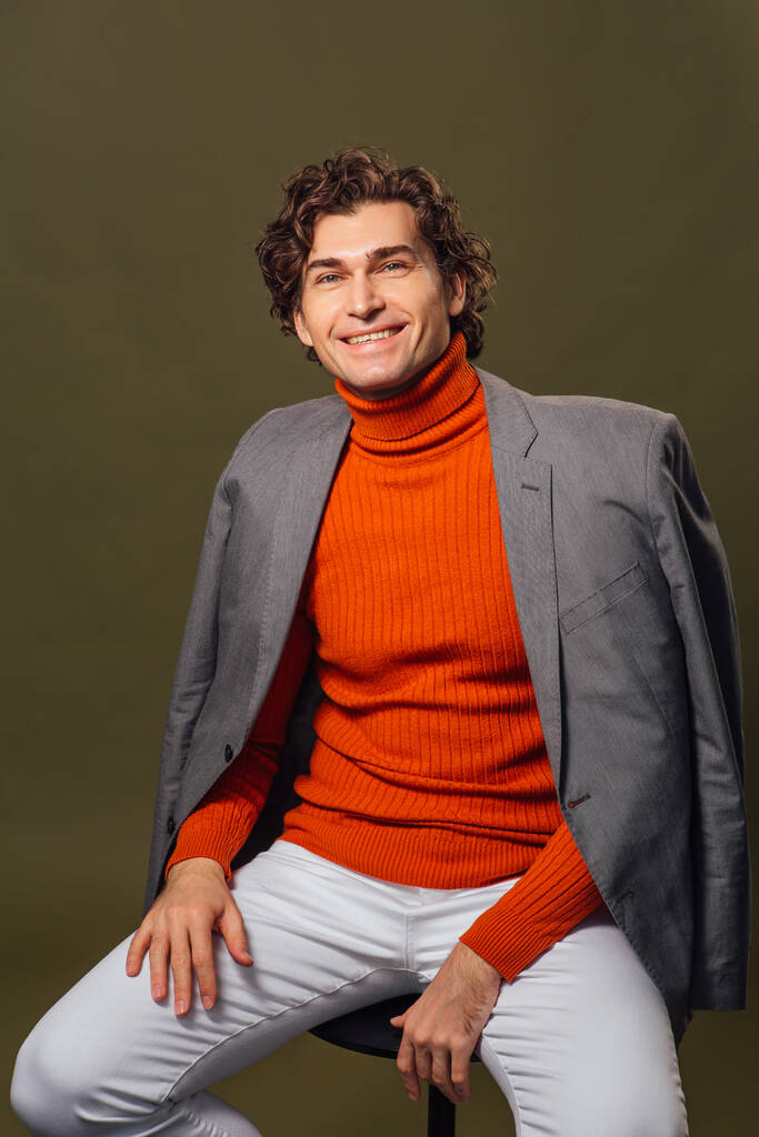 Porträt eines großen, gut aussehenden Mannes mit orangefarbenem Rollkragen, grauer Jacke und weißer Jeans, der auf grünem Hintergrund posiert - Foto, Bild