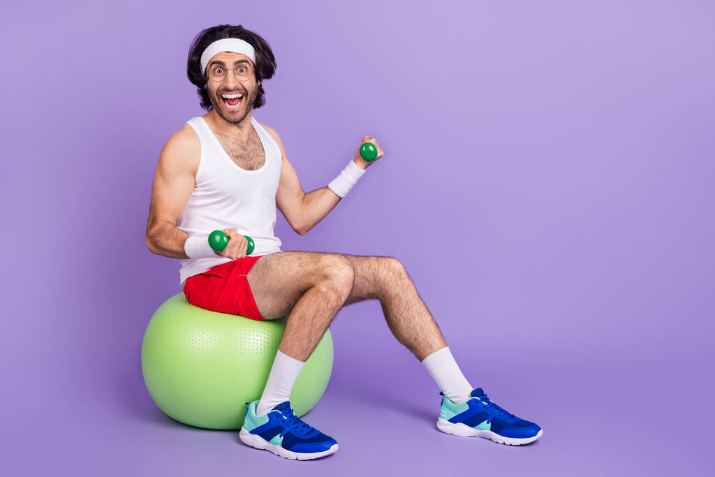 Full size photo of funky happy young man hold dumbbells sit green fit ball smile isolated on purple color background - Photo, Image