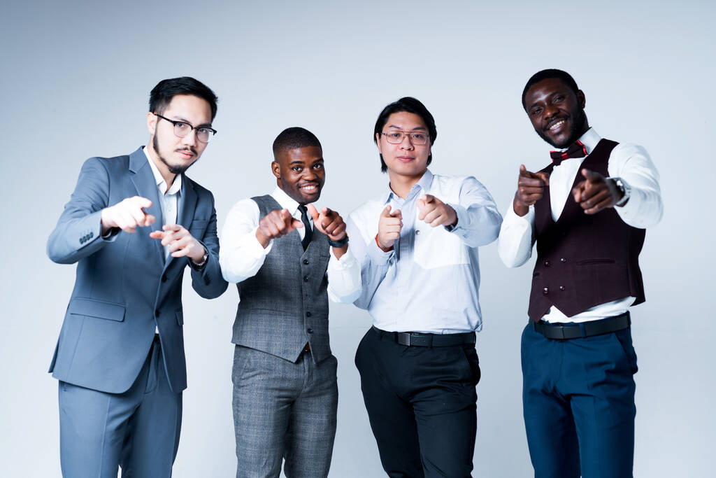 Afrikkalainen amerikkalainen ja aasialainen liikemiehet puvuissa hymyillen ja osoittaen kameraan seisoessaan kevyellä taustalla - Valokuva, kuva