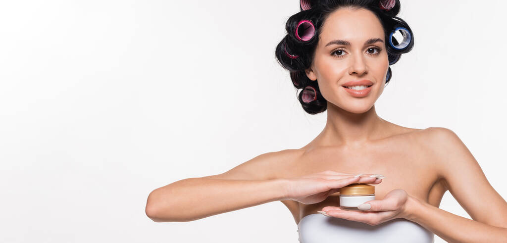 smiling young woman in white bra with curlers on hair holding cream container in hands isolated on white, banner - Photo, Image