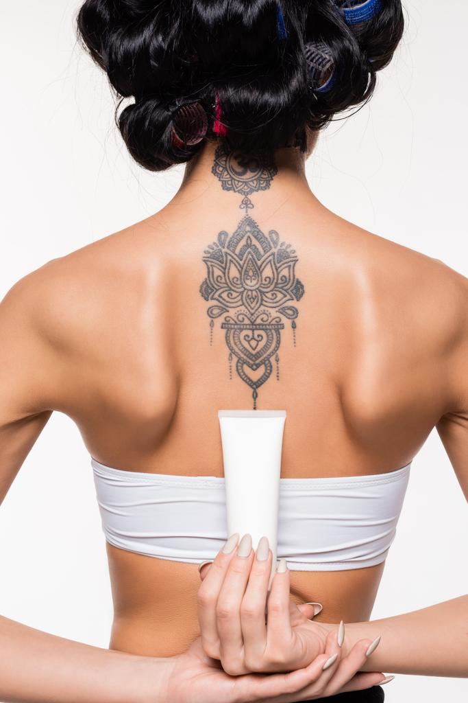 zpět pohled mladé ženy v natáčky s tetováním drží krém trubice izolované na bílém - Fotografie, Obrázek
