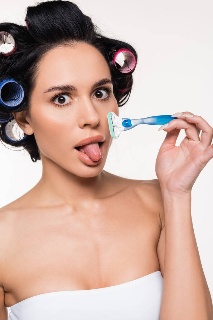 young woman in curlers and top grimacing with sticking out tongue and holding razor near face isolated on white - Photo, Image