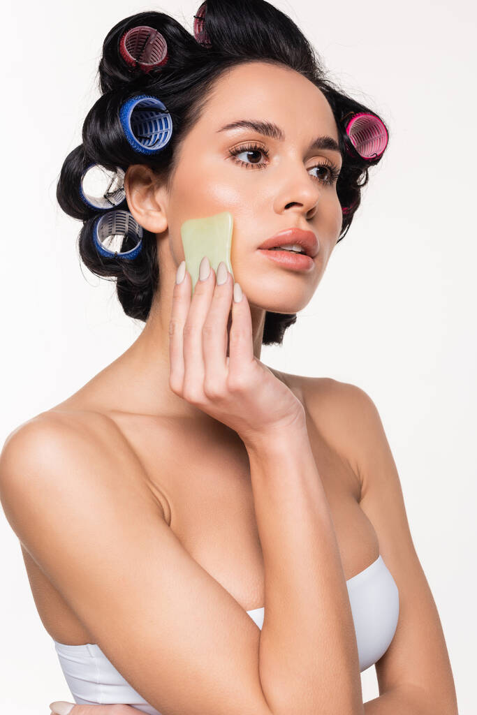 ernstige jonge vrouw in krulspelden en top maken gezichtsmassage met gua sha geïsoleerd op wit - Foto, afbeelding