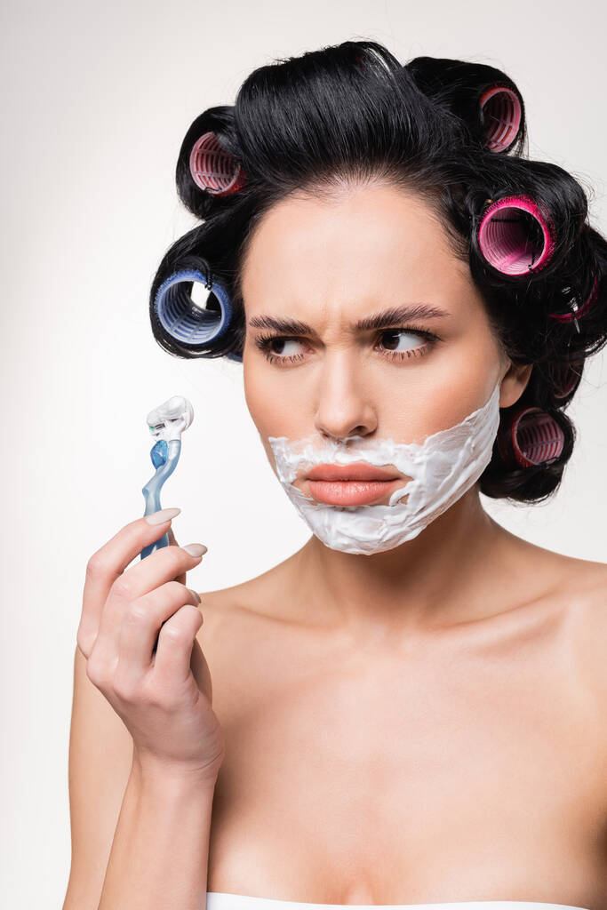 confusa giovane donna in bigodini tenuta rasoio vicino al viso con crema da barba isolata su bianco - Foto, immagini