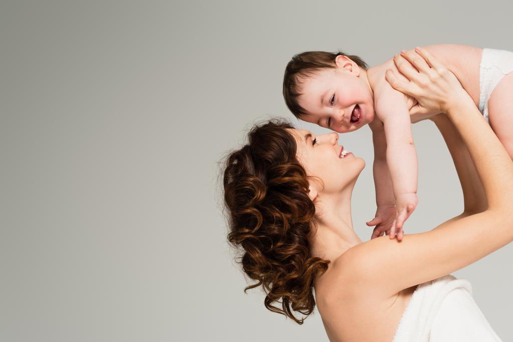 alegre madre con hombros desnudos sosteniendo en brazos bebé niño aislado en gris - Foto, imagen