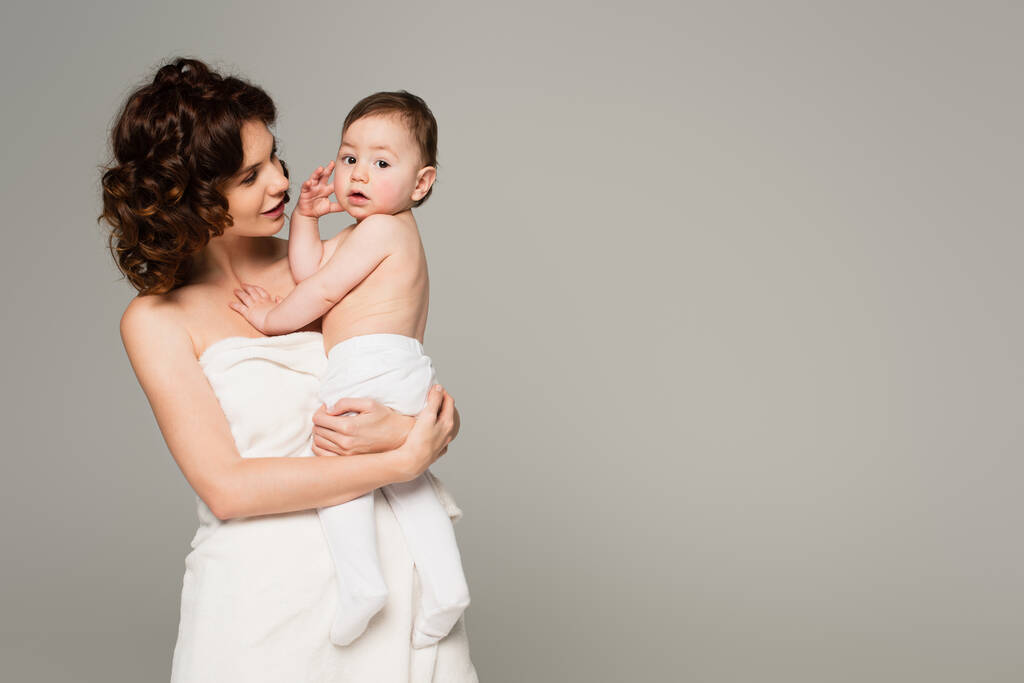 lockige Mutter in Handtuch gehüllt, Sohn im Arm isoliert auf grau  - Foto, Bild