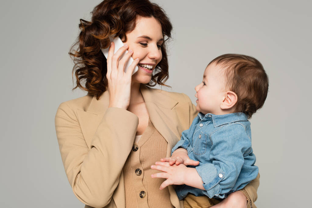 幸せな母親は携帯電話で話し、灰色で隔離された腕の赤ちゃんの男の子を抱えて  - 写真・画像