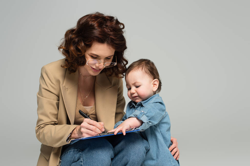 sonriente mujer de negocios en gafas escribiendo en portapapeles cerca de niño pequeño hijo aislado en gris - Foto, imagen