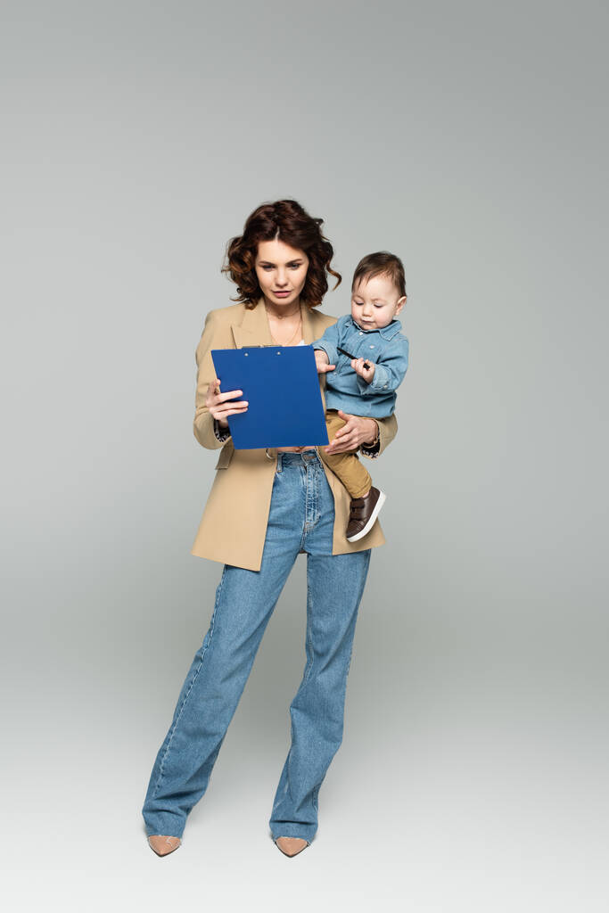 volledige lengte van zakenvrouw in blazer houden klembord en baby jongen in armen op grijs - Foto, afbeelding