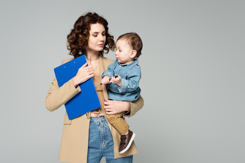 göndör üzletasszony blézerben kezében írótáblával és kisfiúval a karjában elszigetelve a szürkén - Fotó, kép