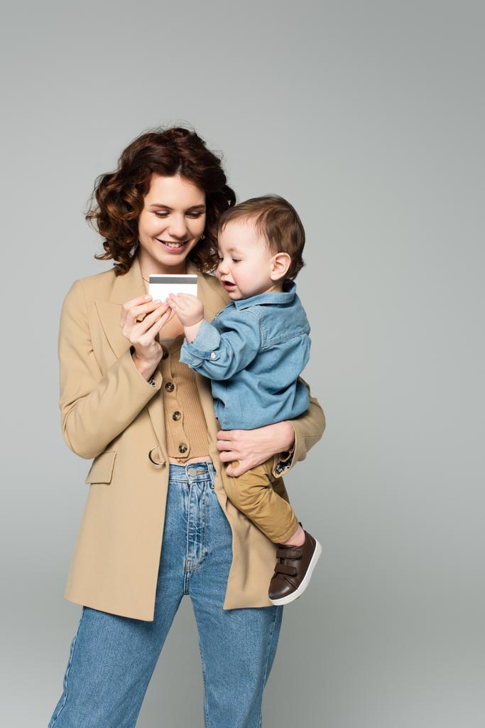 happy woman holding in arms toddler son and credit card isolated on grey - Photo, Image