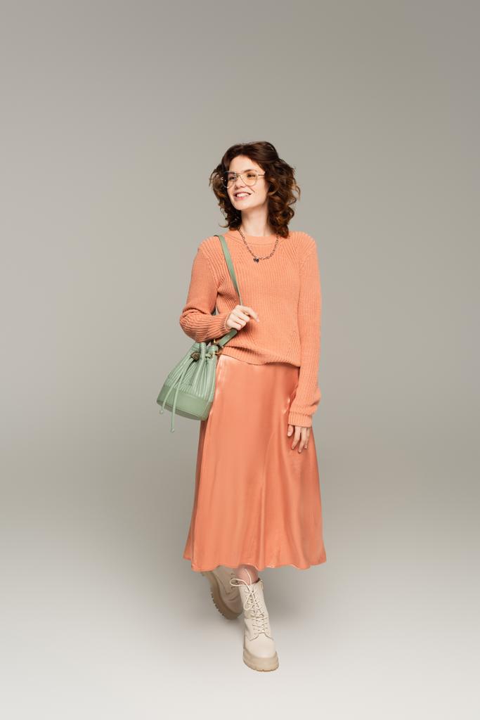 full length of stylish woman in glasses, sweater and silk skirt holding leather strap of bag on grey  - Photo, Image