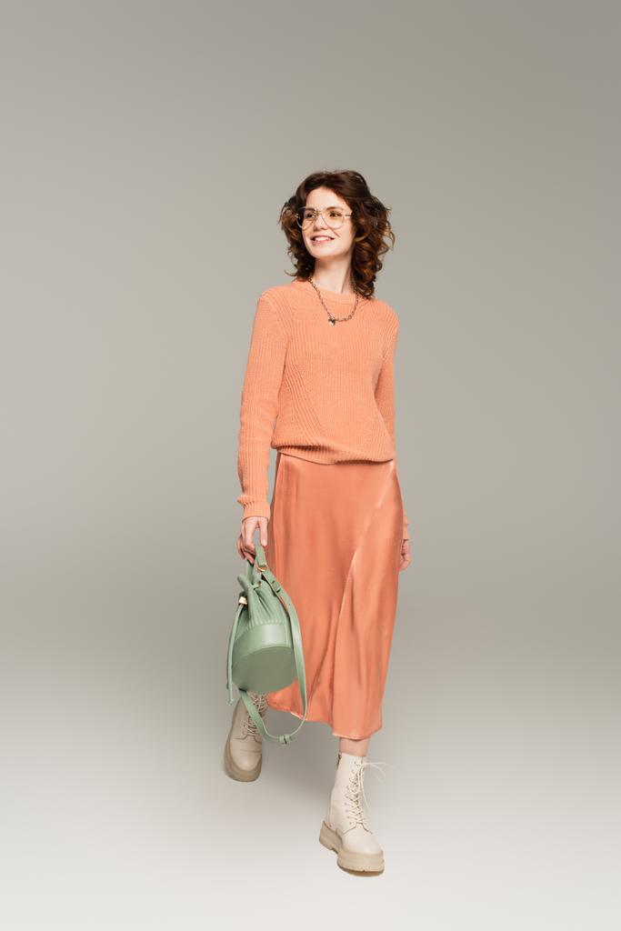 volledige lengte van gelukkig vrouw in bril en perzik trui holding bag tijdens het lopen op grijs  - Foto, afbeelding