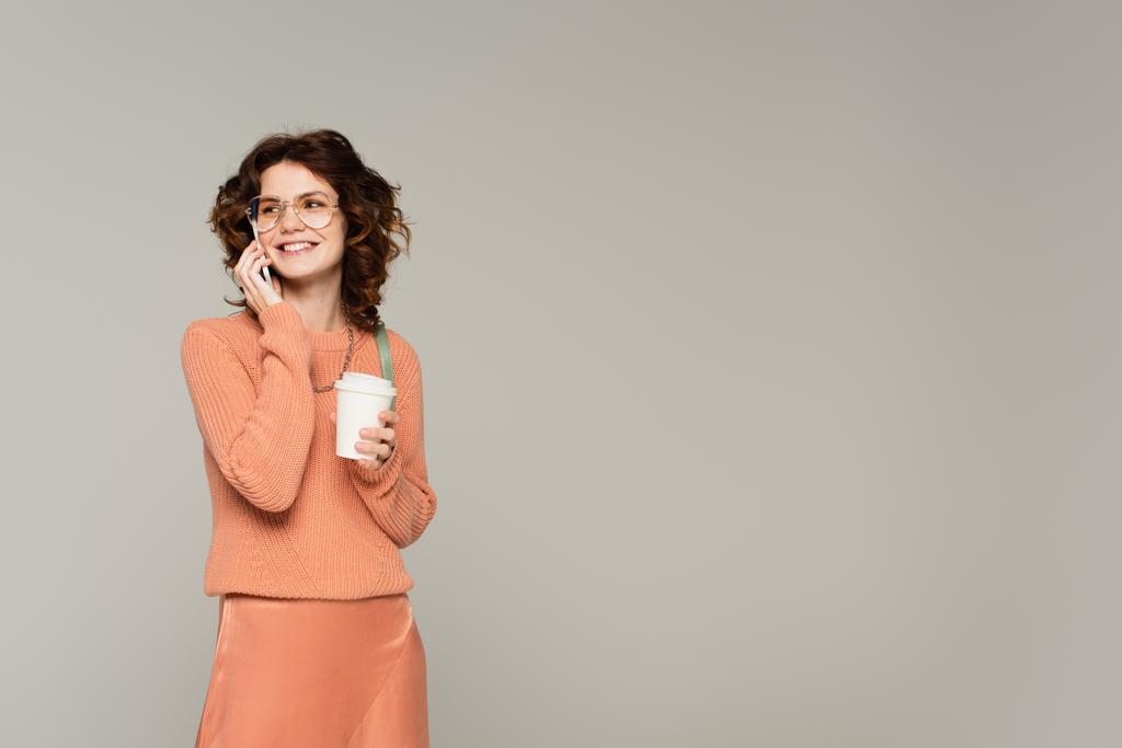boldog nő pulóverben kezében kávé menni, miközben beszél mobiltelefon elszigetelt szürke  - Fotó, kép
