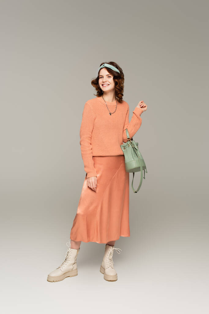 полная длина счастливой женщины в повязке, свитере и атласной юбке, стоящей на сером  - Фото, изображение