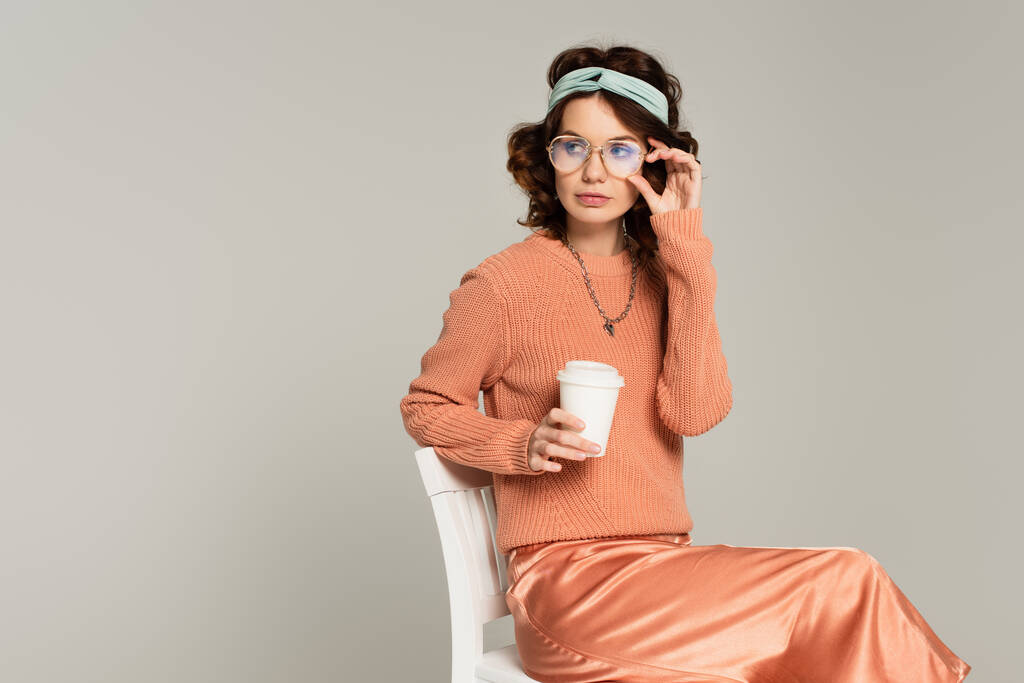 kudrnatá žena nastavení brýle a drží papírový šálek, zatímco sedí na židli izolované na šedé  - Fotografie, Obrázek