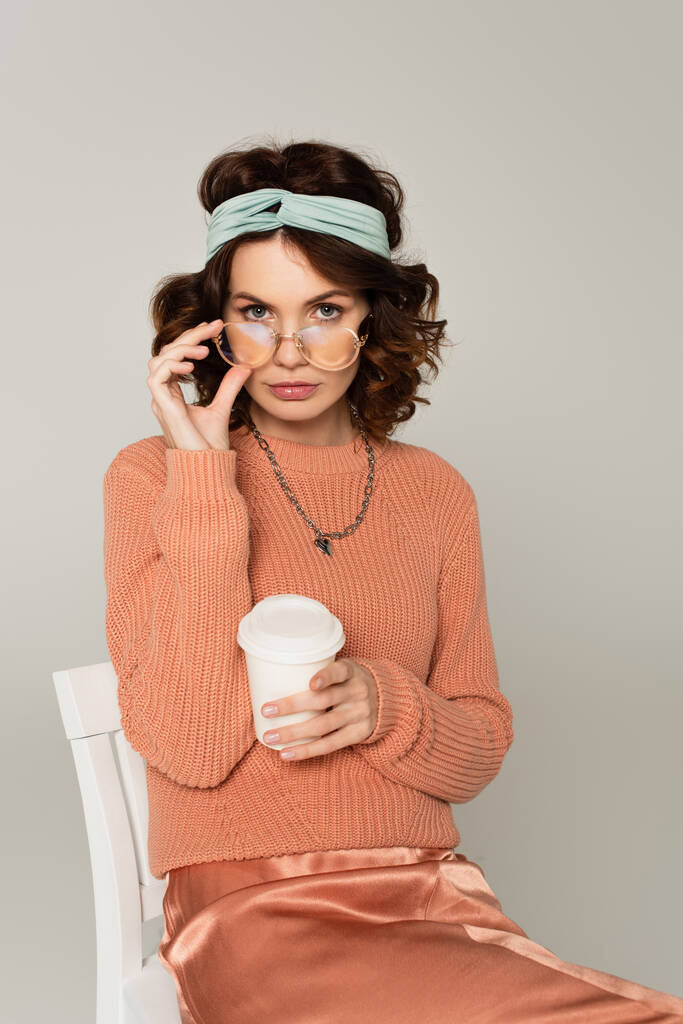 stylish woman adjusting glasses and holding paper cup while sitting on chair isolated on grey  - Photo, Image