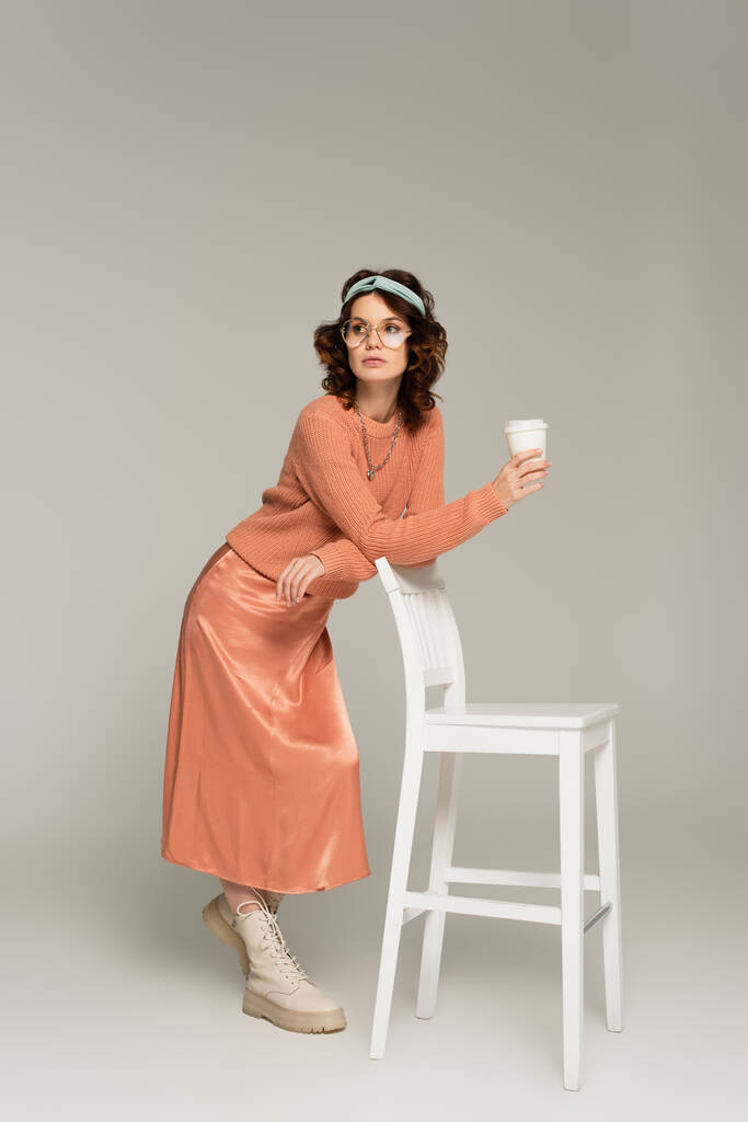 pełna długość kręconych kobiet w okularach i opasce opartej na krześle trzymając papierowy kubek na szarości  - Zdjęcie, obraz