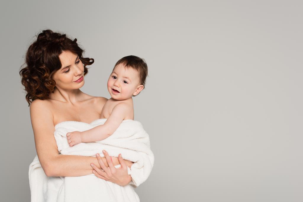 fürsorgliche Mutter mit nackten Schultern, die Kleinkind-Sohn in Handtuch gewickelt isoliert auf grau hält - Foto, Bild