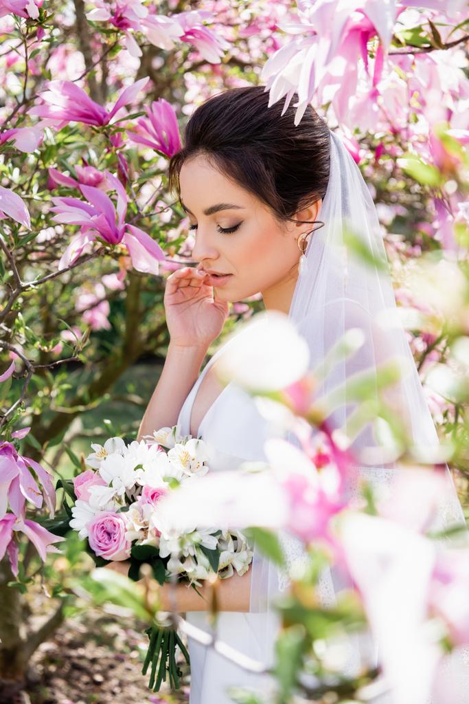 Jeune mariée voilée debout près d'un magnolia en fleurs  - Photo, image