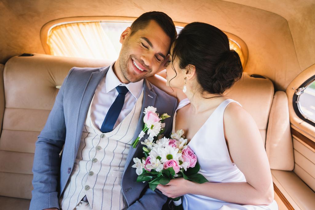 Mariée en robe tenant bouquet près du marié souriant en voiture vintage  - Photo, image