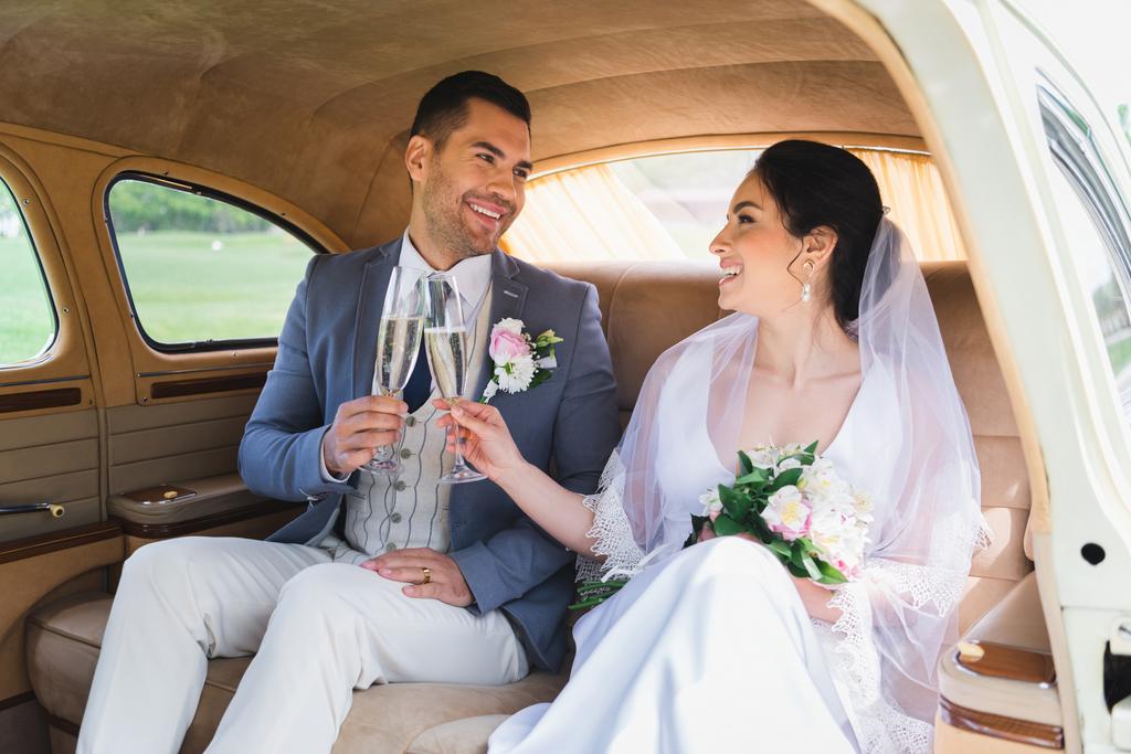 Happy bride and groom clinking with champagne in vintage car  - Photo, Image