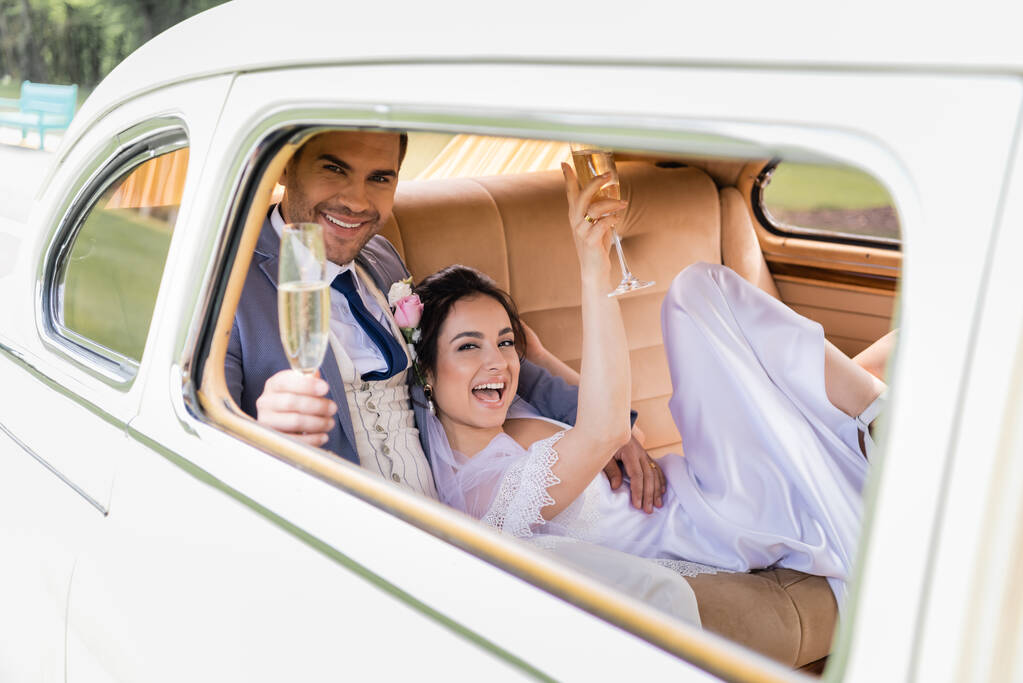 Fröhlicher Bräutigam hält verschwommenes Glas Champagner neben Braut im Retro-Auto  - Foto, Bild