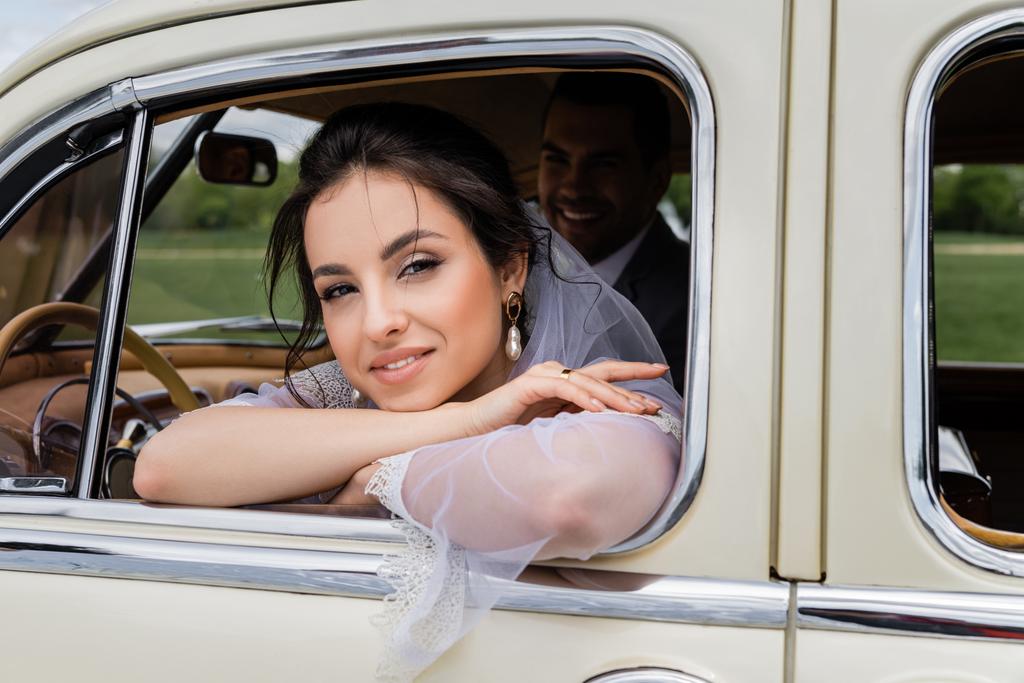 Cheerful bride in white veil looking at camera near window of retro car - Photo, Image