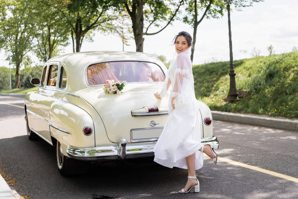 Ευτυχισμένη νύφη στέκεται κοντά μπουκέτο σε vintage αυτοκίνητο στο δρόμο  - Φωτογραφία, εικόνα
