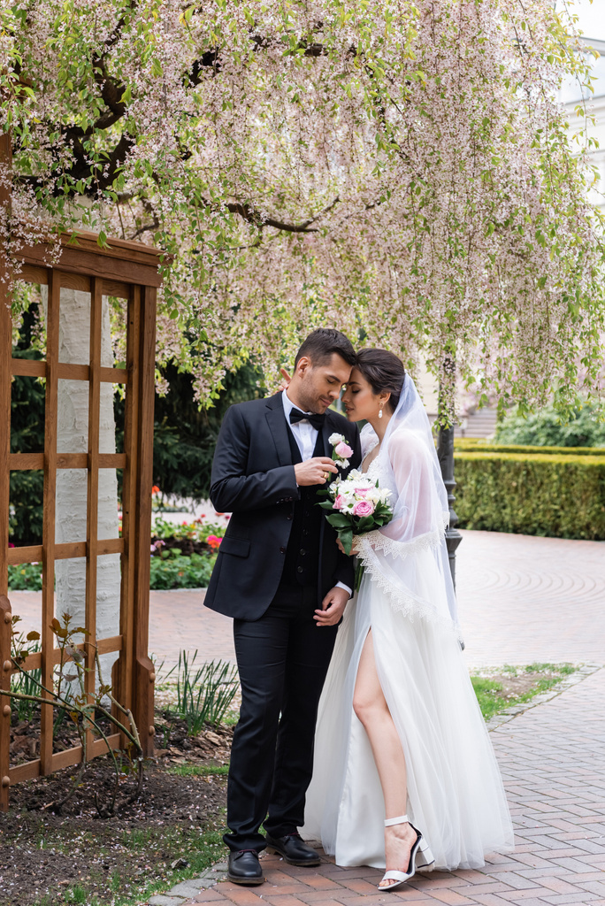 Boční pohled na mladé novomanžele stojící v blízkosti kvetoucího stromu v parku  - Fotografie, Obrázek
