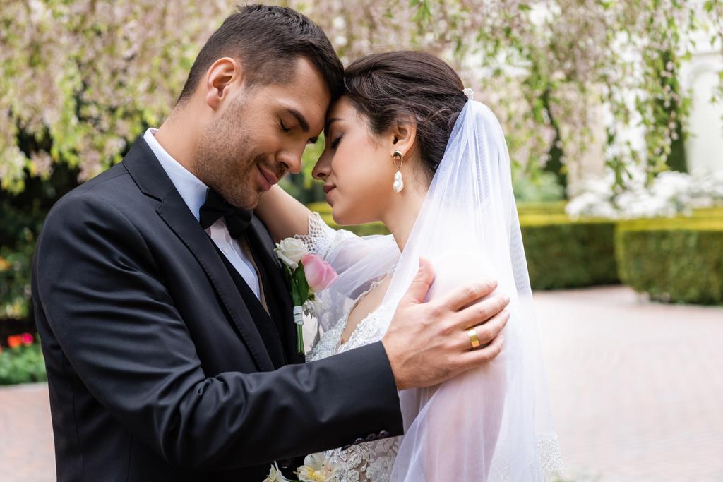 Vista laterale dello sposo che abbraccia la giovane sposa con gli occhi chiusi nel parco  - Foto, immagini