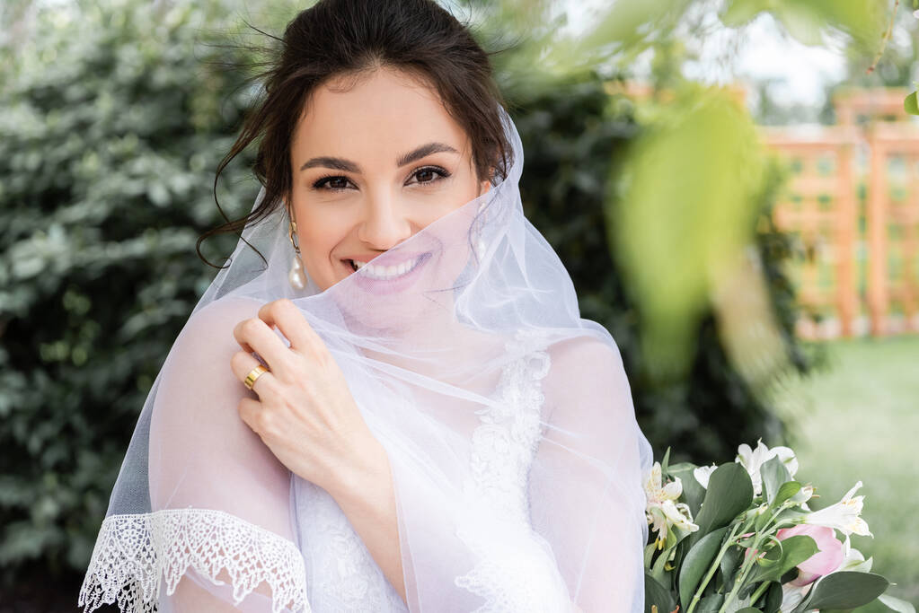 Mariée joyeuse tenant voile blanc près du bouquet à l'extérieur  - Photo, image