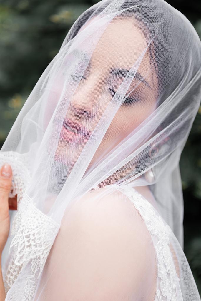 Улыбающаяся невеста в белой вуали стоит с закрытыми глазами  - Фото, изображение